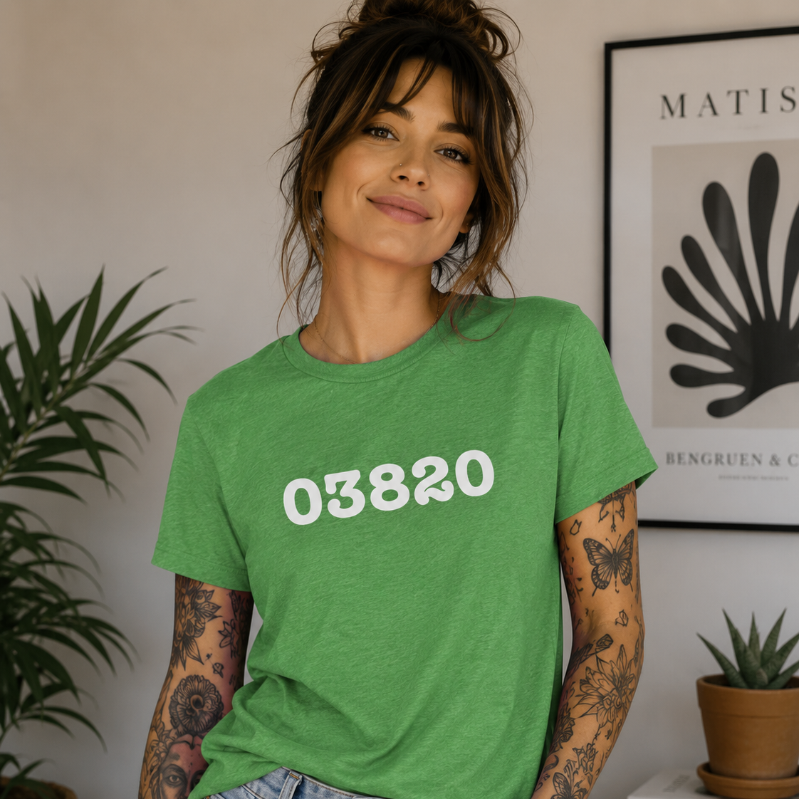 Woman wearing a green t-shirt with '03820' printed on it, standing in a room with plants and decor.