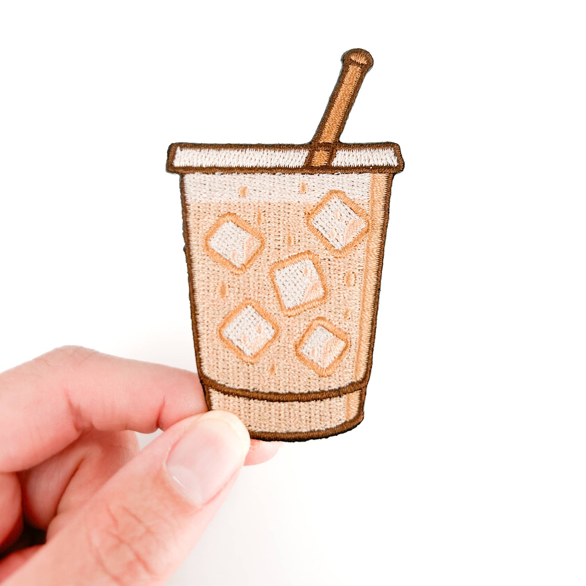 Small embroidered patch of a coffee cup with ice and a straw held by a hand on a white background