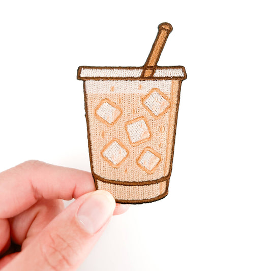 Small embroidered patch of a coffee cup with ice and a straw held by a hand on a white background