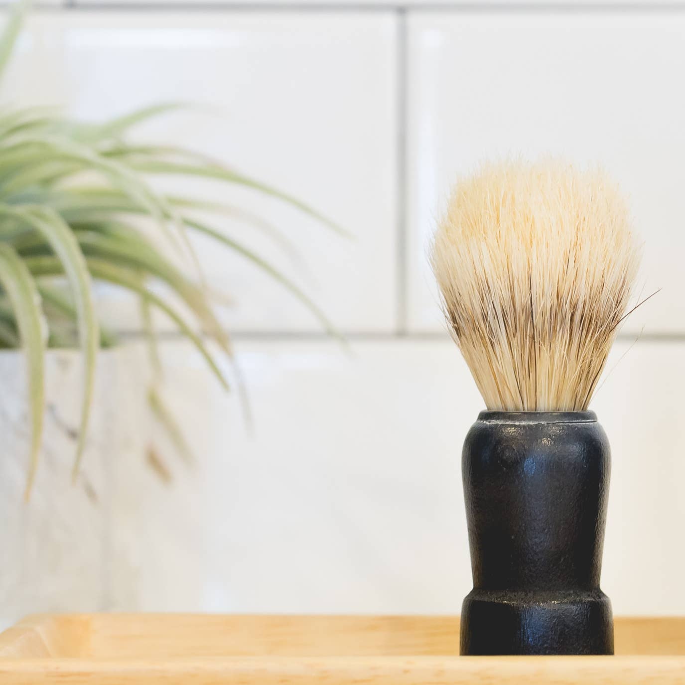 Shaving brush with black handle and natural bristles on a wooden surface with a plant in the background