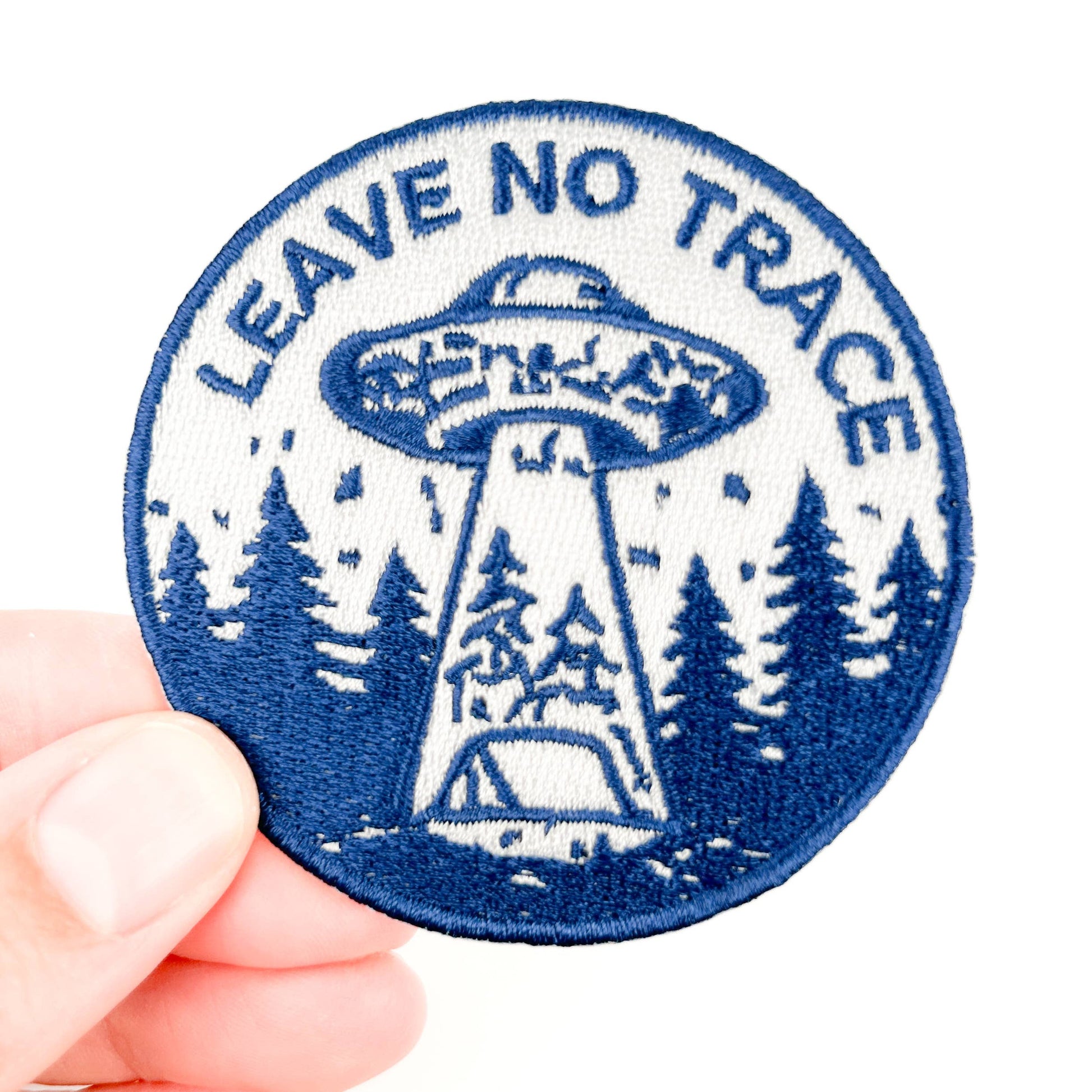 Blue 'Leave No Trace' patch with UFO design held by a hand on a white background