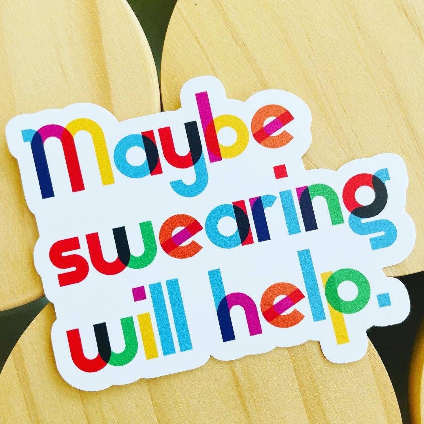 Colorful sticker with text 'maybe swearing will help.' on a wooden background