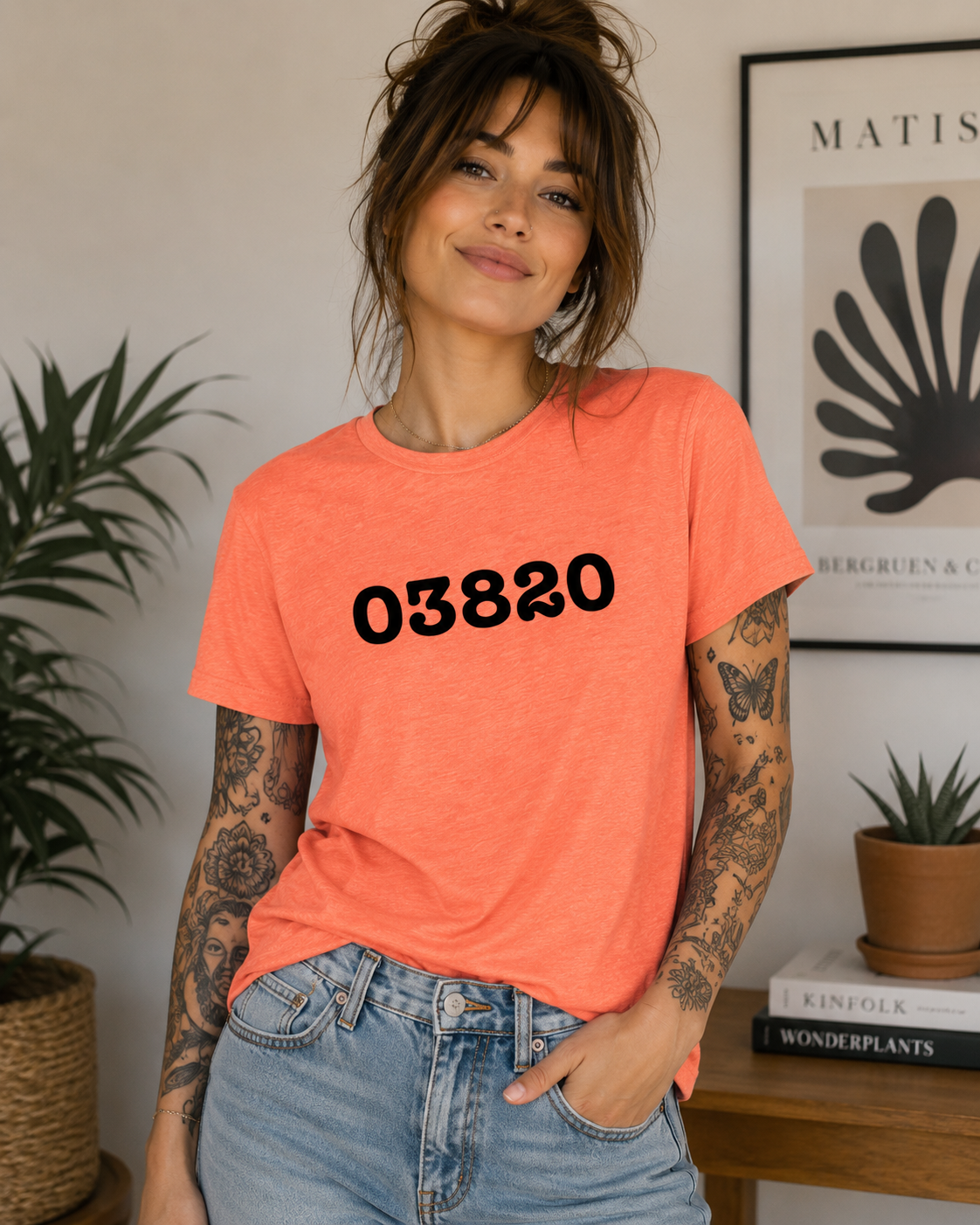 Woman wearing an orange t-shirt with '03820' printed on it, standing in a room with plants and a framed picture.
