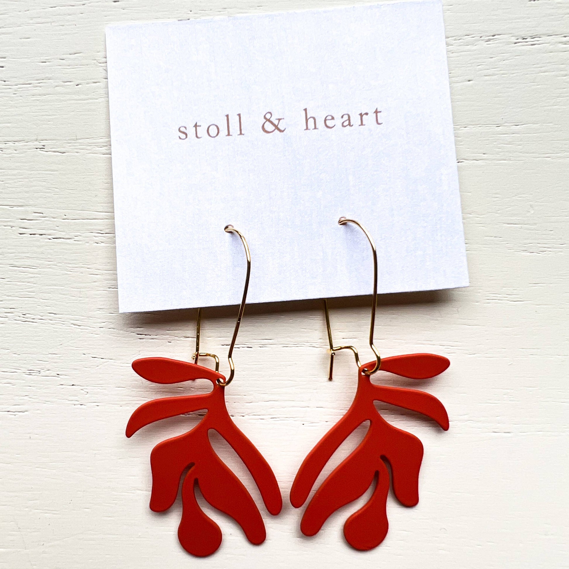 Red earrings on a white card with 'stoll & heart' branding on a light wooden surface.