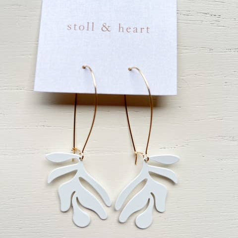 White leaf-shaped earrings on gold hooks with a 'stoll & heart' card on a light wooden surface.