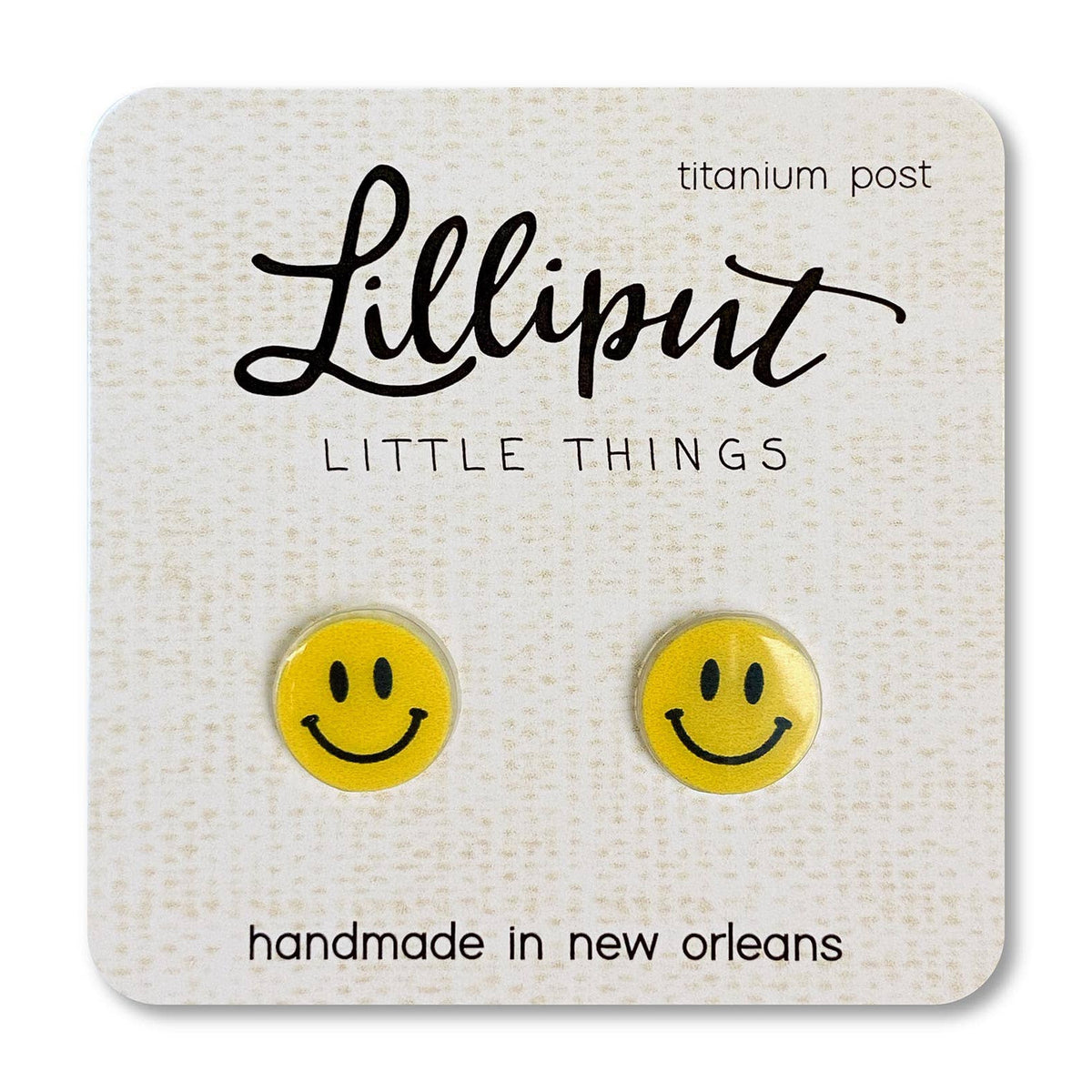 A pair of yellow smiley face stud earrings on a white Lilliput brand display card, placed on a wooden surface.